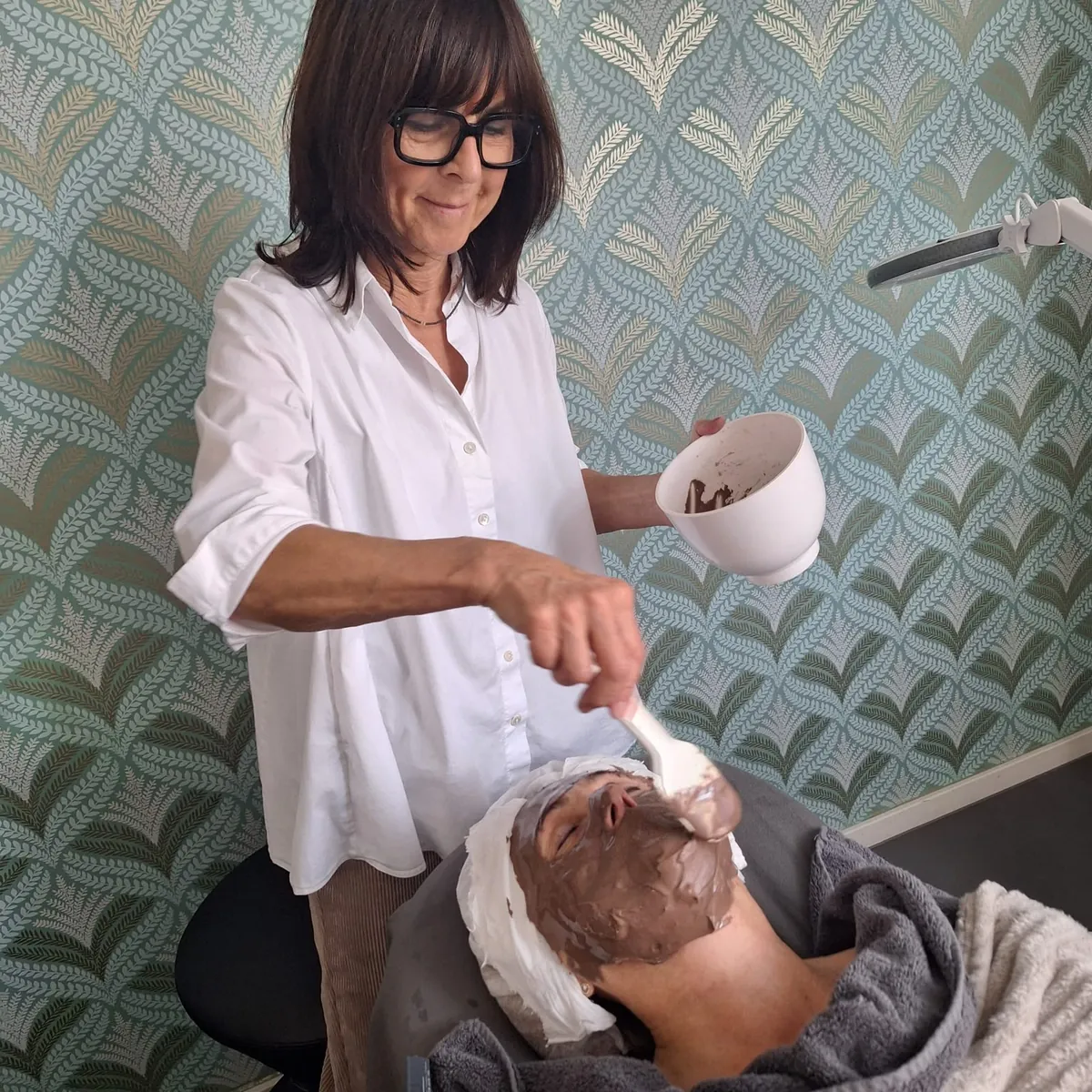 Sabine Eisebraun trägt bei einer Kundin die Kaffeemaske der Glow Behandlung auf
